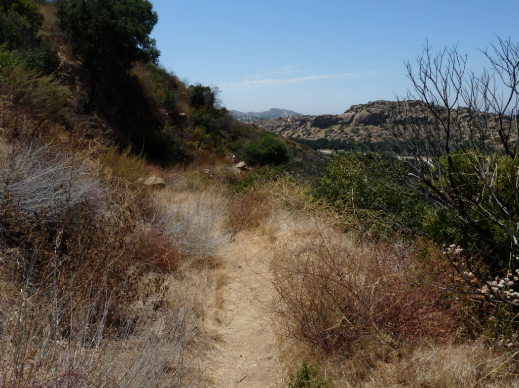 A dirt trail winds through dry grasses and shrubs on Stagecoach Road, viewed westward with hills and a clear blue sky in the distance. Photographed in 2010 by Mary K. Maki, M.A., RPA, Archaeologist.