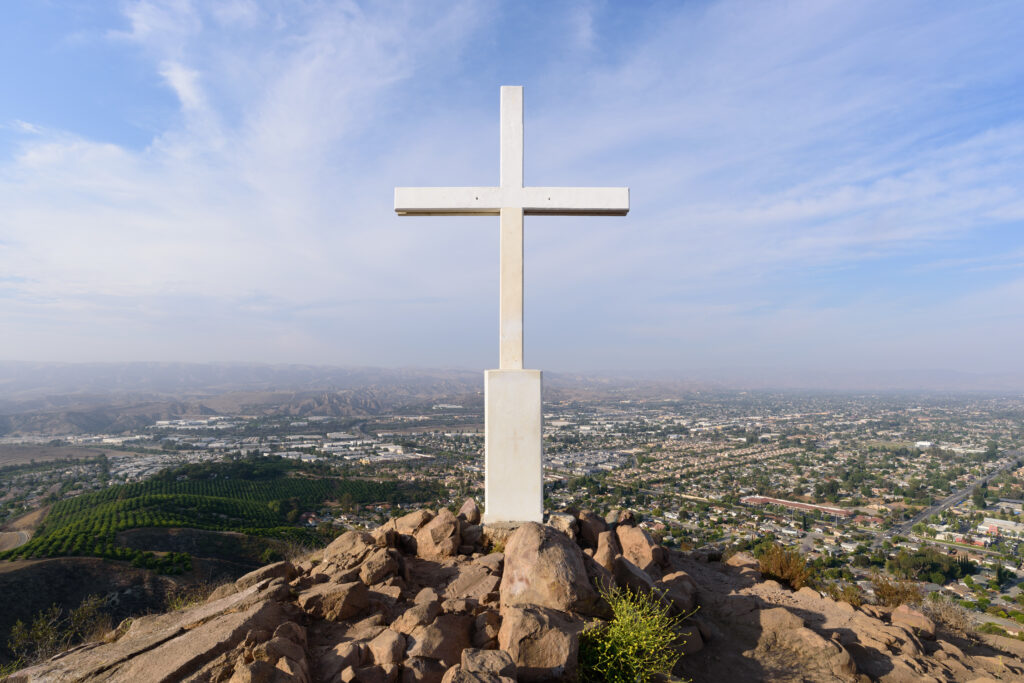 A white cross atop Mt. McCoy overlooks Simi Valley, with a sprawling urban area and distant hills under a partly cloudy sky.