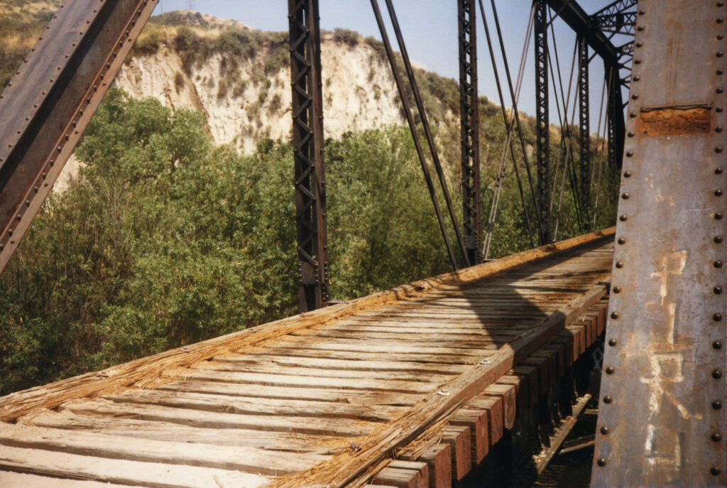The Piru Train Bridge, circa 1980s, features a weathered wooden deck with metal trusses and rusted beams, set against rocky cliffs and greenery.