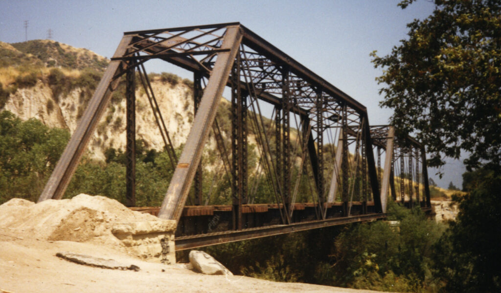 Steel truss train bridge spanning a valley with rocky hills and greenery, featuring a sand pile in the foreground; circa 1980s.