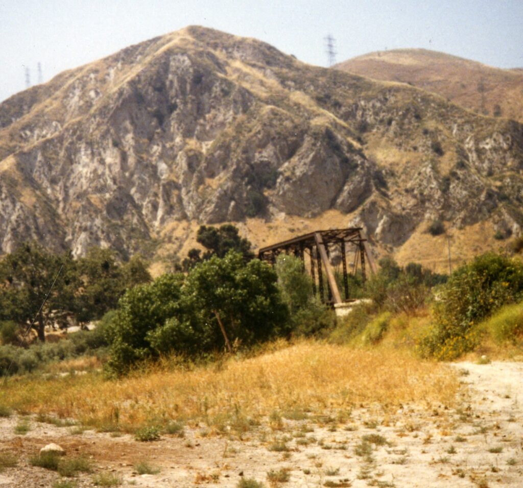 An abandoned steel train bridge structure stands in a dry, grassy field with rocky mountains in the background, circa 1980s.