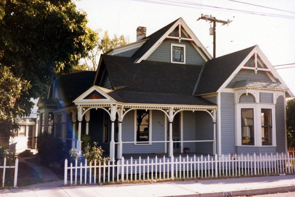 Rice House, a light blue Victorian home with white trim, wraparound porch, and white picket fence, circa 1980s. Address number 928 visible on the sidewalk.
