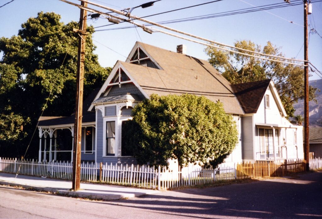 A blue and white Victorian house with a white picket fence, mature trees, and utility poles, identified as the Rice House from the 1980s.