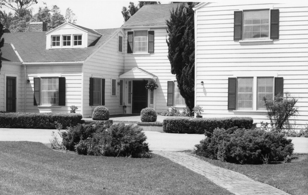 Black-and-white photograph of the Richard Bard House, circa 1980s, featuring a white clapboard house with dark shutters, a dormer window, and a landscaped garden with trimmed bushes and a brick pathway.