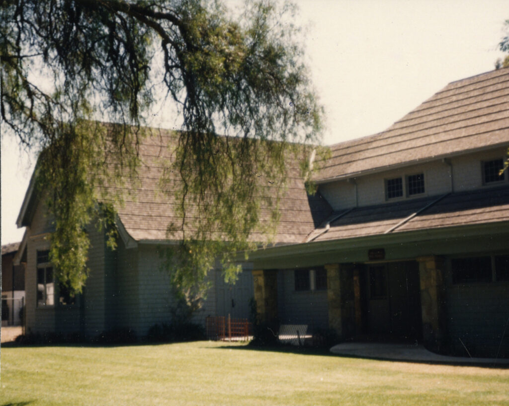 Light gray Santa Paula Ebell Club building from the 1980s, featuring a shingled roof, large willow tree, lawn, and covered porch with garage doors.