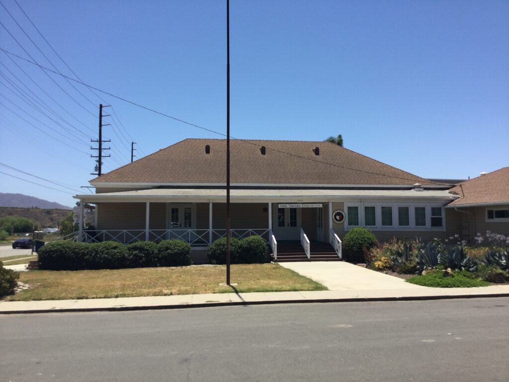 Single-story beige clubhouse with brown roof and white trim, featuring a covered porch with white railings and steps to the entrance; visible signage above the door reads "Somis Thursday Clubhouse".