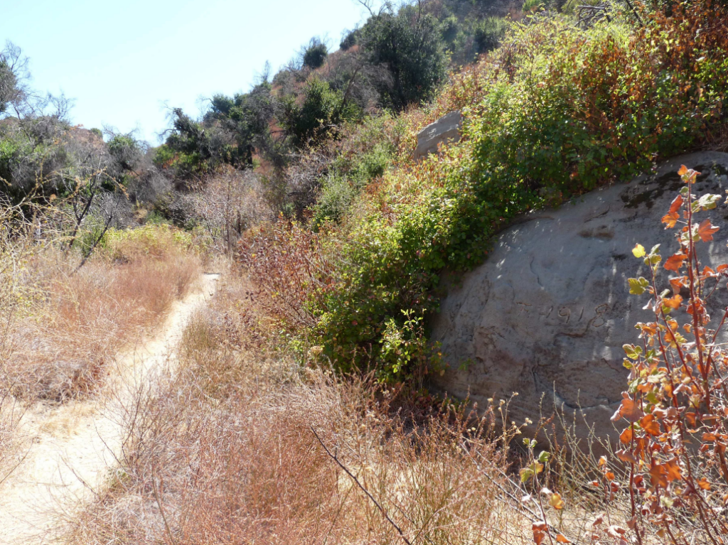 A dirt path winds east on Stagecoach Road in 2010, bordered by dry vegetation and green shrubs, with a large rock on the right featuring the carved date 1918 under a clear sky.