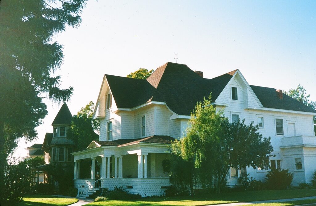 White Victorian house with dark shingled roof, wraparound porch, and surrounding trees on a sunny day. Captioned Underwood House, 1985.