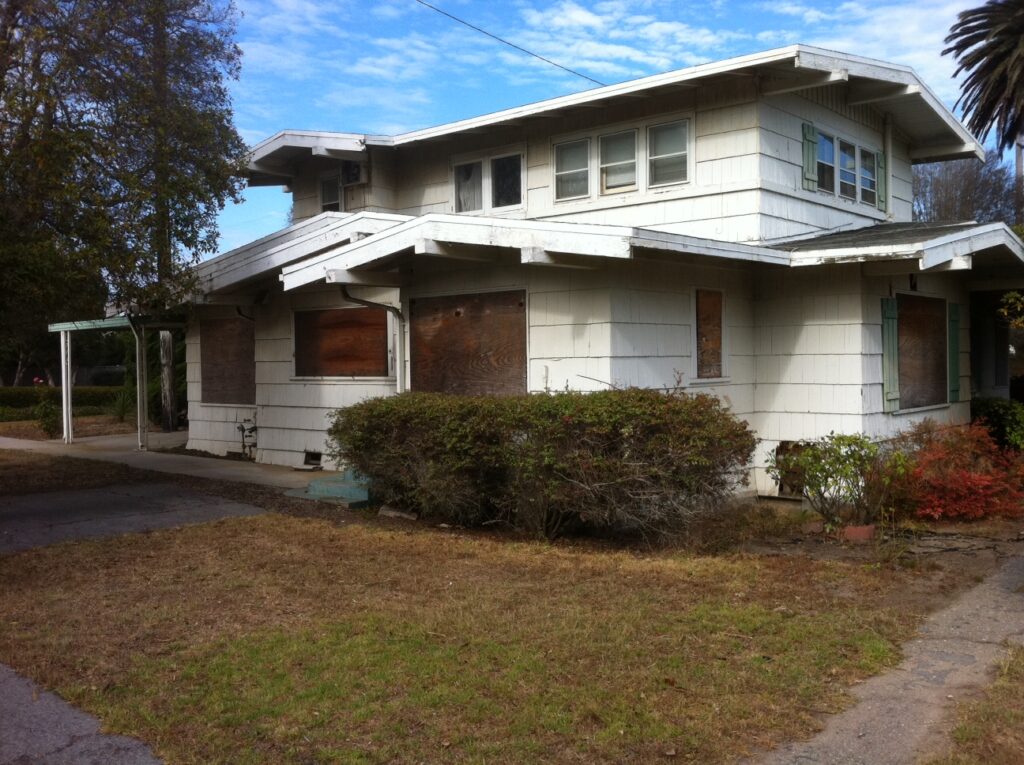 Vacant two-story house with boarded windows, overgrown bushes, and dry lawn in disrepair.