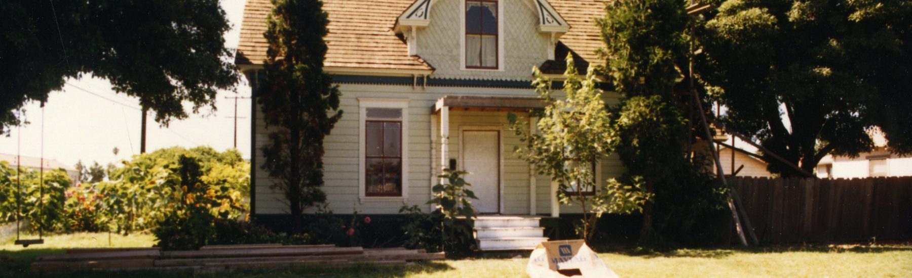 Light yellow Victorian house with green trim, gabled roof, and porch, surrounded by a wooden picket fence, trees, and garden, identified as the Gerberding/Moranda House from the 1980s.