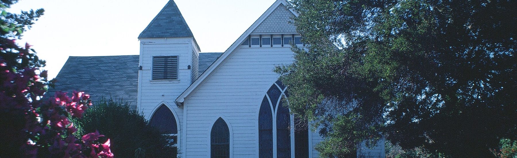 White wooden church with Gothic windows and steeple, surrounded by trees and pink flowers, photographed in 1981.