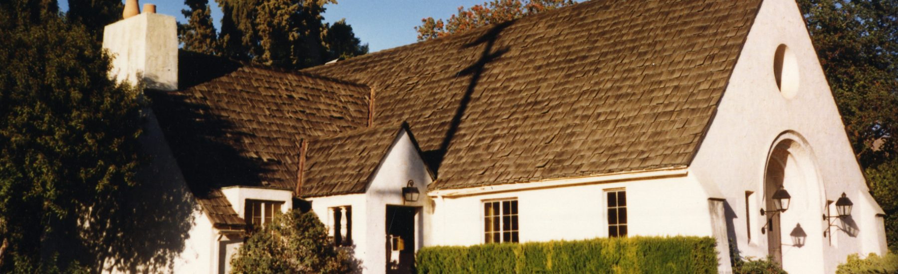 White church building with dark shingled roof, arched doorway, and chimney, surrounded by trees and greenery. Built in 1985.
