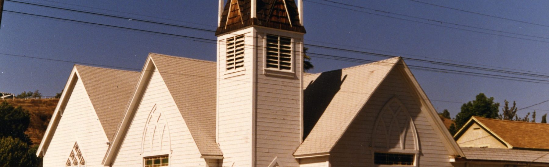 White wooden Methodist church with a steeple and Gothic windows, photographed in 1985. Features central entrance, multiple windows, and an adjacent structure under a clear blue sky.