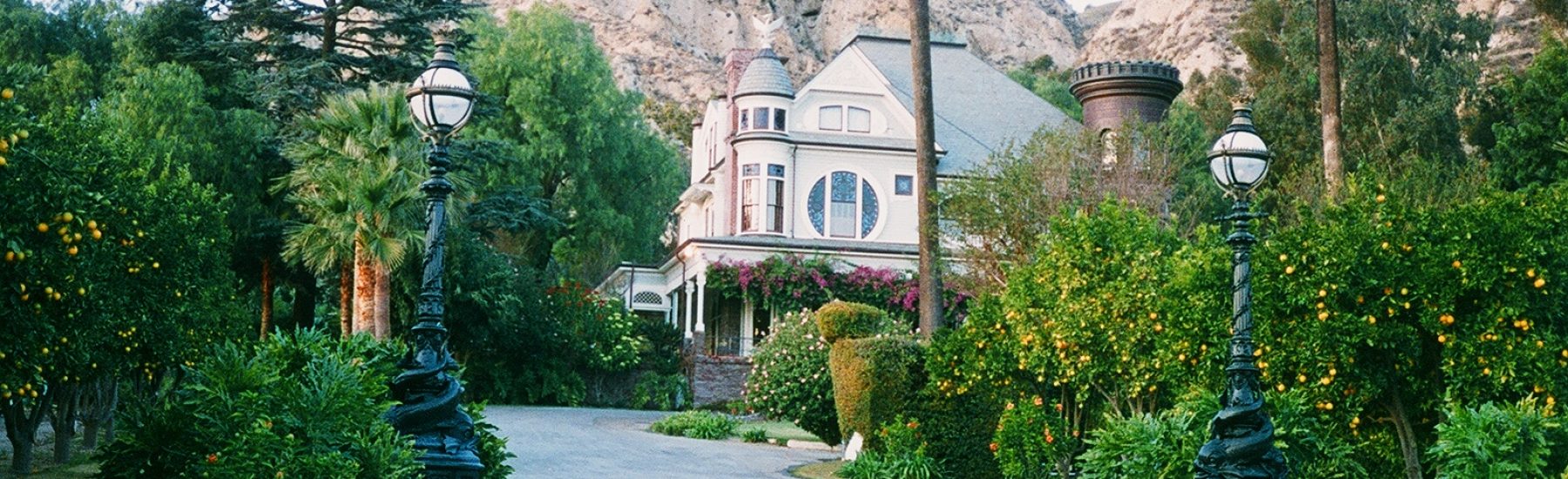 Historic mansion with ornate light fixtures, surrounded by lush greenery and citrus trees, nestled against a rocky hillside.