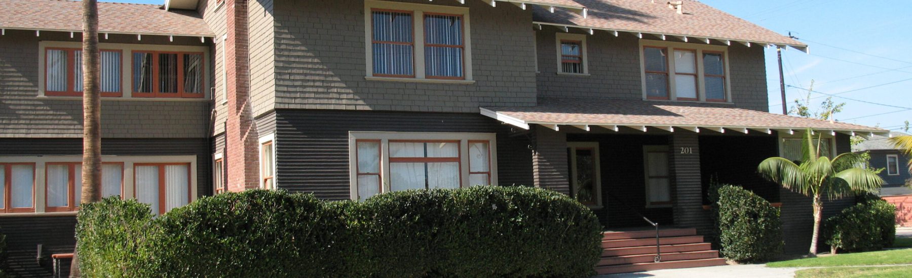 Achille Levy House, a two-story residence with brick and siding exterior, red-trimmed windows, covered porch with steps, and address '201'. Features landscaped lawn with hedges and palm trees under a clear blue sky.