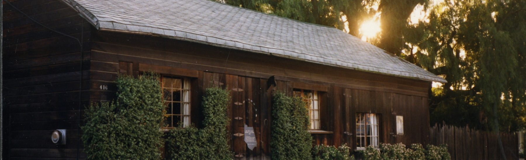 Wooden Artists' Barn from the 1980s with a gray shingled roof, brick chimney, and greenery around windows. Orange flowers and the address number 416 are visible in front of the building.