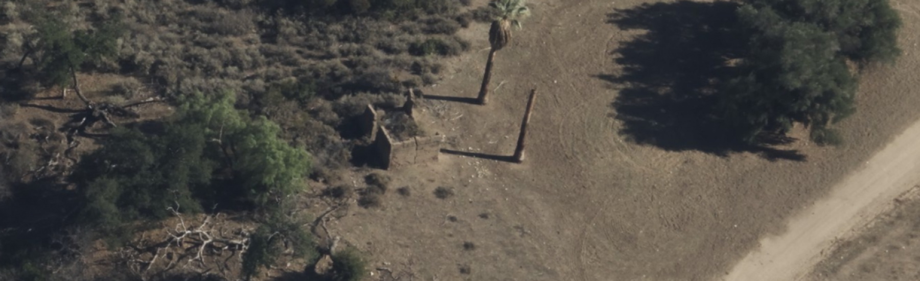 Aerial view of Arundell Adobe ruins with stone walls, a palm tree, and surrounding desert vegetation near a curving dirt road.