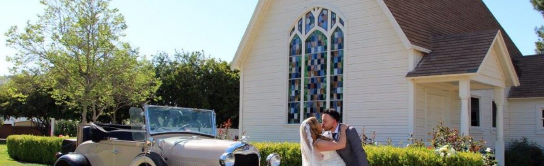 A bride in a white gown and groom in a gray suit embrace on a grassy lawn in front of Saint Rose of Lima Catholic Church, with a vintage tan and black convertible (license plate '5LUR480') parked nearby. The church features a colorful stained glass window and white siding.