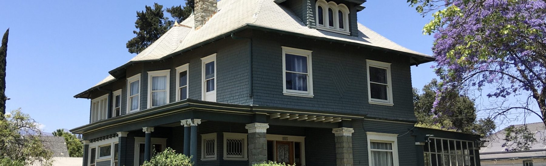 Dark gray Victorian house with white trim, stone chimney, and porch columns. Purple jacaranda tree and green lawn in front. Charles Collins Teague House, 2021, photographed by Craig Baker.
