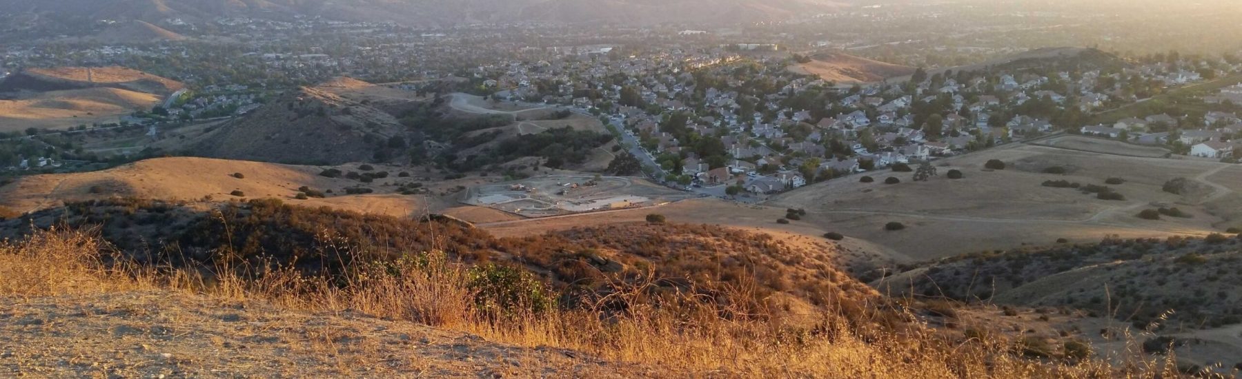 Panoramic view from Chumash Trail lookout, featuring dry grassy hills, suburban neighborhoods, and distant mountains illuminated by warm sunlight.