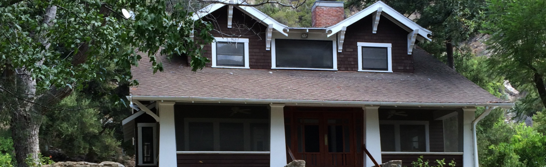 Brown shingle house with white trim and stone foundation, featuring a wooden front porch with stairs, surrounded by trees and a rocky hillside.