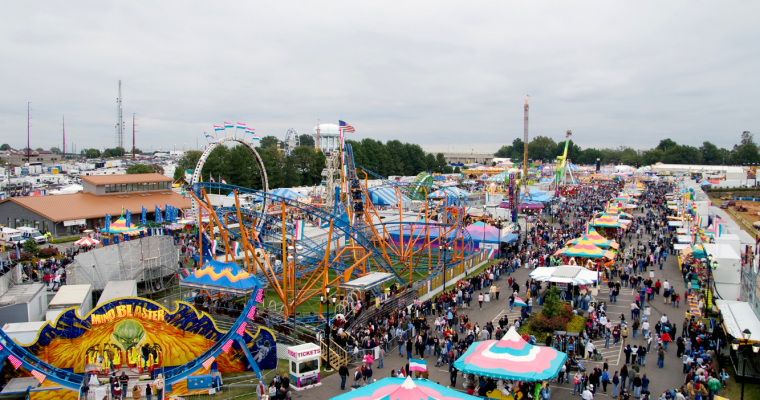 Aerial view of a local fairground with various rides, including a large roller coaster, tents with colorful tops, and a crowd of people walking through the fair.