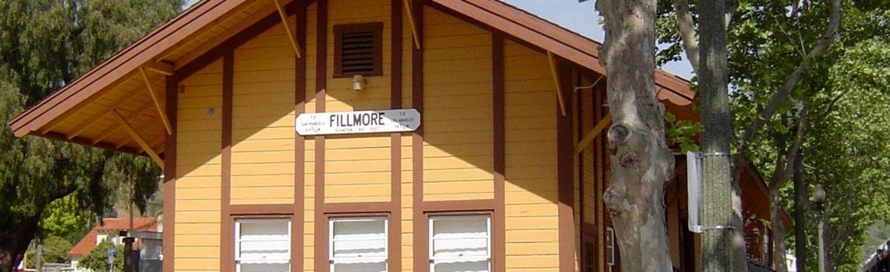 Yellow and brown wooden railroad depot with a "FILLMORE" sign, three windows with white shutters, and a covered porch, situated along railroad tracks with trees in the foreground.