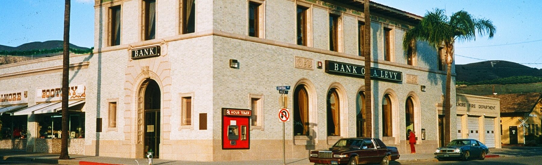 1986 Fillmore State Bank building with "BANK" and "BANK OF A LEVY" signs, a red "24 HOUR TELLER" ATM, palm trees, parked cars, and a nearby fire department building.
