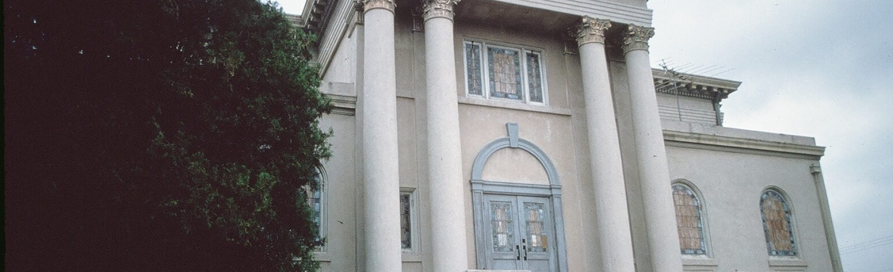 Former Simi Valley Community Methodist Church building with four columns, arched blue door, red steps, stained glass windows, and a large tree on the left.