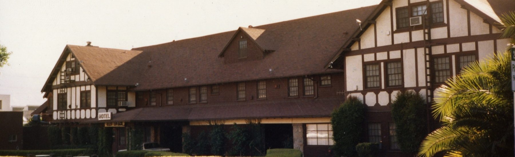 Tudor-style Glen Tavern Hotel from 1985 with white and brown timber framing, a brown roof, and a visible "HOTEL" sign. Manicured green hedges and palm trees frame the building's exterior.