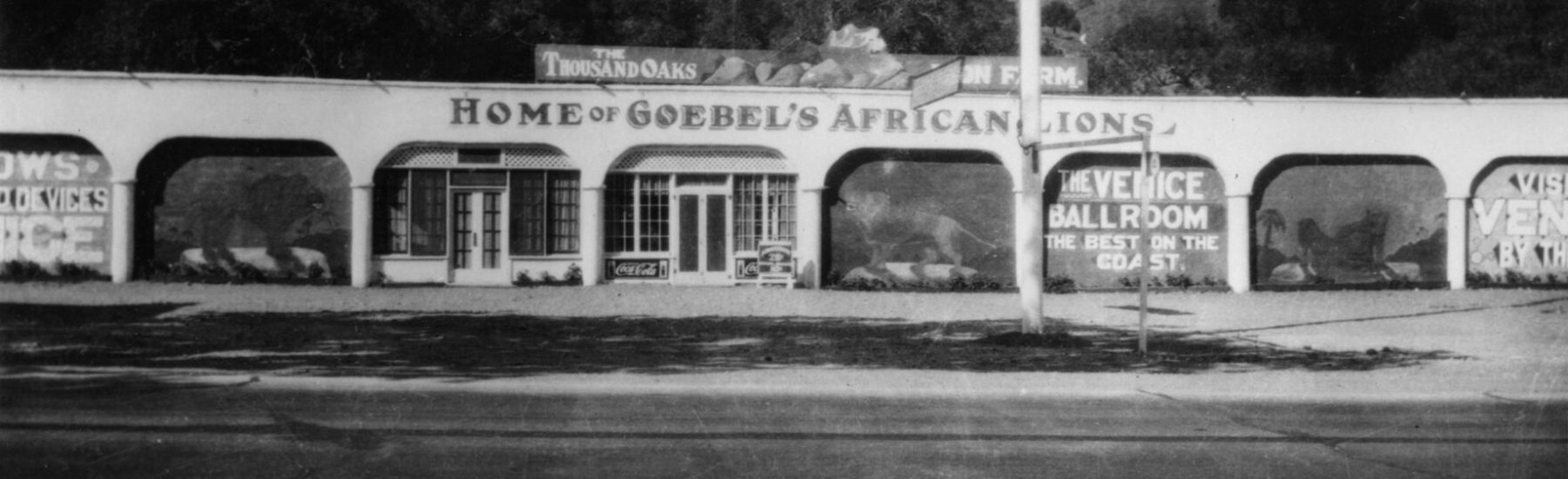 Black-and-white photograph of Goebel's Lion Farm building, circa 1929, featuring arched openings with visible text including "HOME OF GOEBEL'S AFRICAN LIONS" and "THE VENICE BALLROOM", set against a hilly landscape.