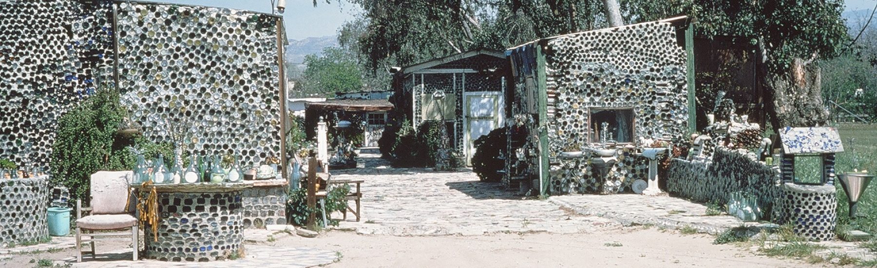 Grandma Prisbrey's 1978 bottle-cap village with structures, furniture, and a dirt path surrounded by trees.