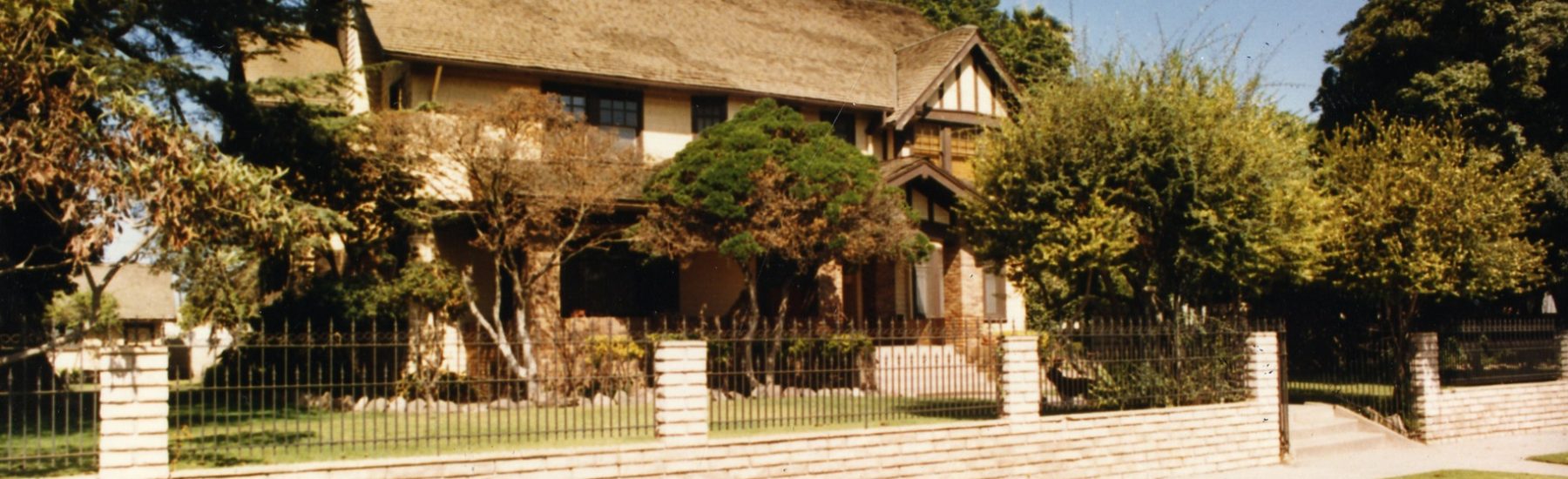 Two-story Henry Levy House with thatched roof, surrounded by trees and a brick fence, circa 1980s.