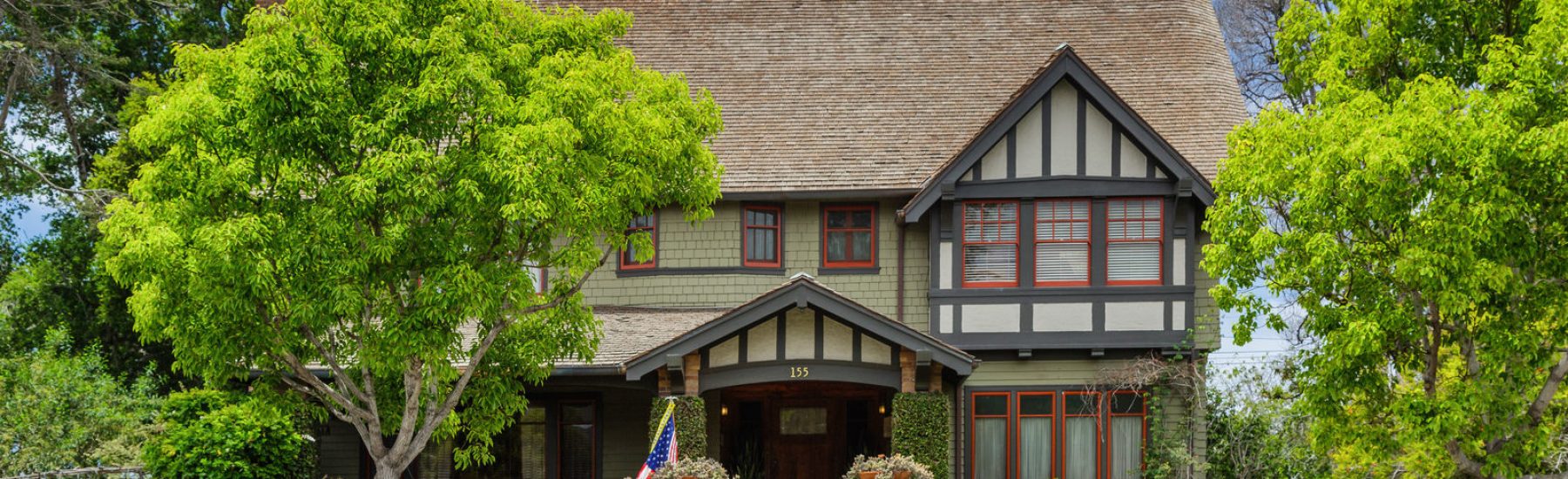 Tudor-style house with green siding, red window frames, and brown shingled roof. Features a front yard with manicured hedges, two trees, an American flag, and address number 155 above the entrance.