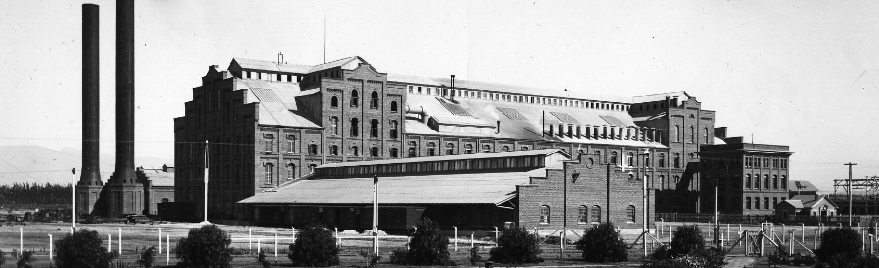 Historical black and white photograph of the Oxnard Sugar Beet Factory, featuring large smokestacks and surrounding grounds.