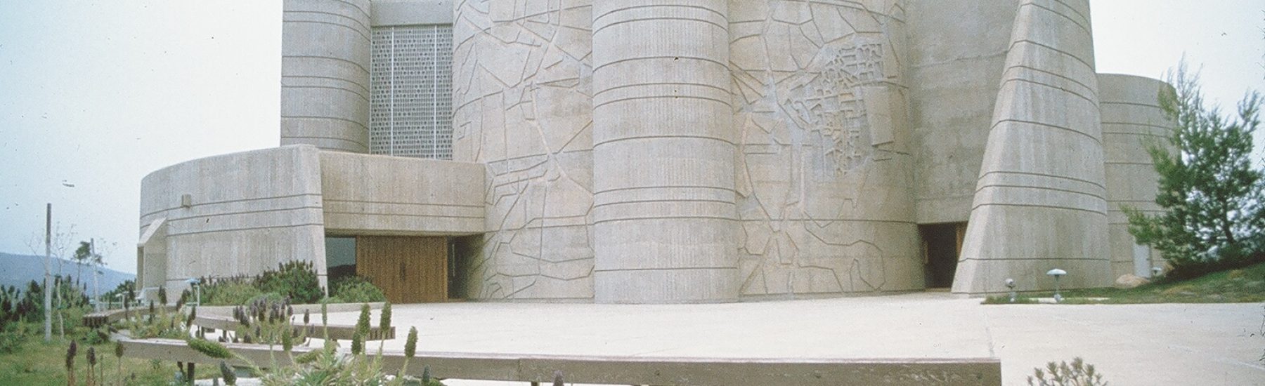 A concrete architectural structure identified as the House of the Book from 1978, featuring cylindrical towers, a curved facade with abstract carvings, and a grassy landscape with sparse vegetation.