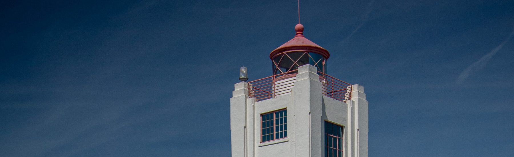 White Hueneme Lighthouse building with a red-domed tower, red-framed windows, and chain-link fence under a clear blue sky. Identified as Hueneme Lighthouse, 2008, with credit to Stephen Schafer.