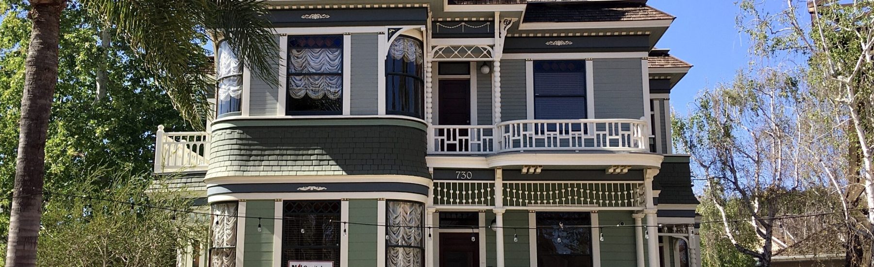A green Victorian-style house with white trim, a turret, balconies, and address '730' displays a 'Mai Capital AVAILABLE' sign.