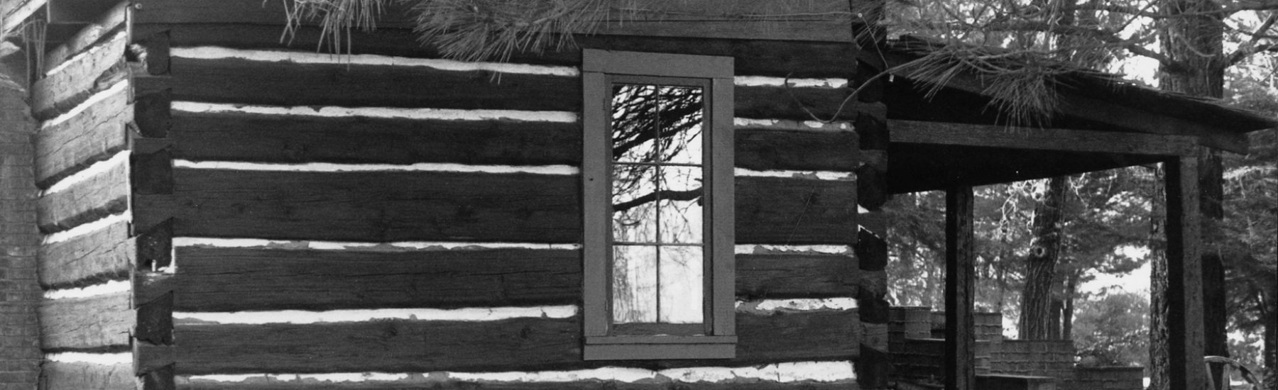 Log cabin with horizontal logs, white chinking, window, stone foundation, and covered porch surrounded by trees. Lathrop Camp, circa 1980s.