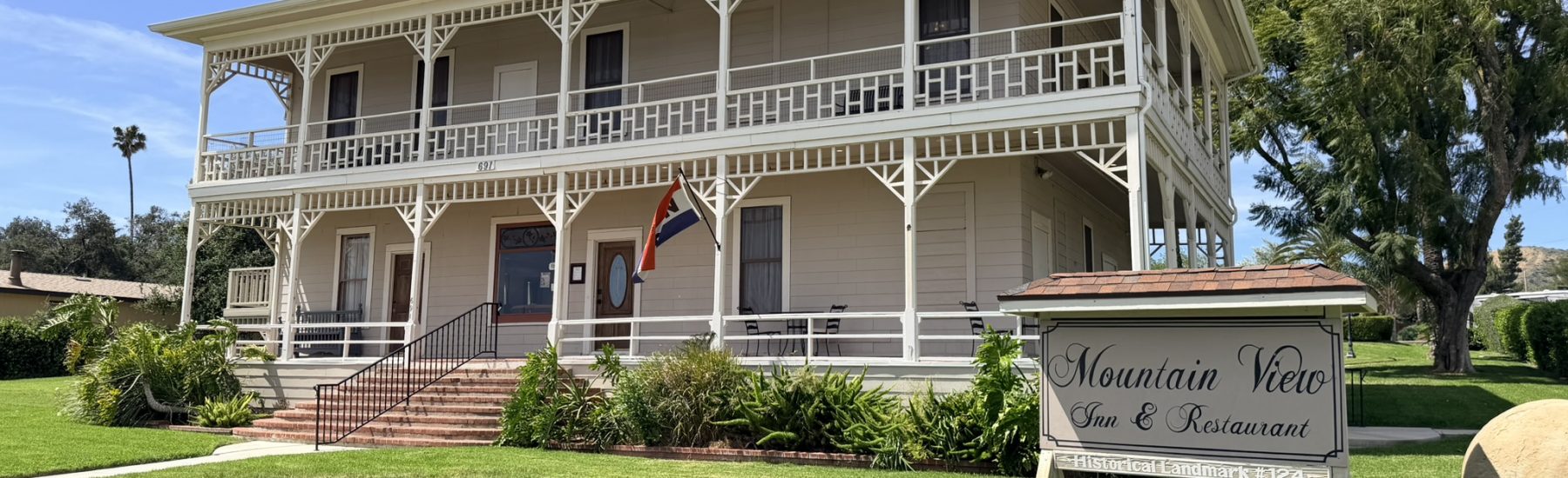 Two-story Victorian-style building with wraparound porch and white railings, featuring a "Mountain View Inn & Restaurant" sign and "Historical Landmark #124" text. Light-colored exterior, flag, and landscaped lawn under a clear blue sky.