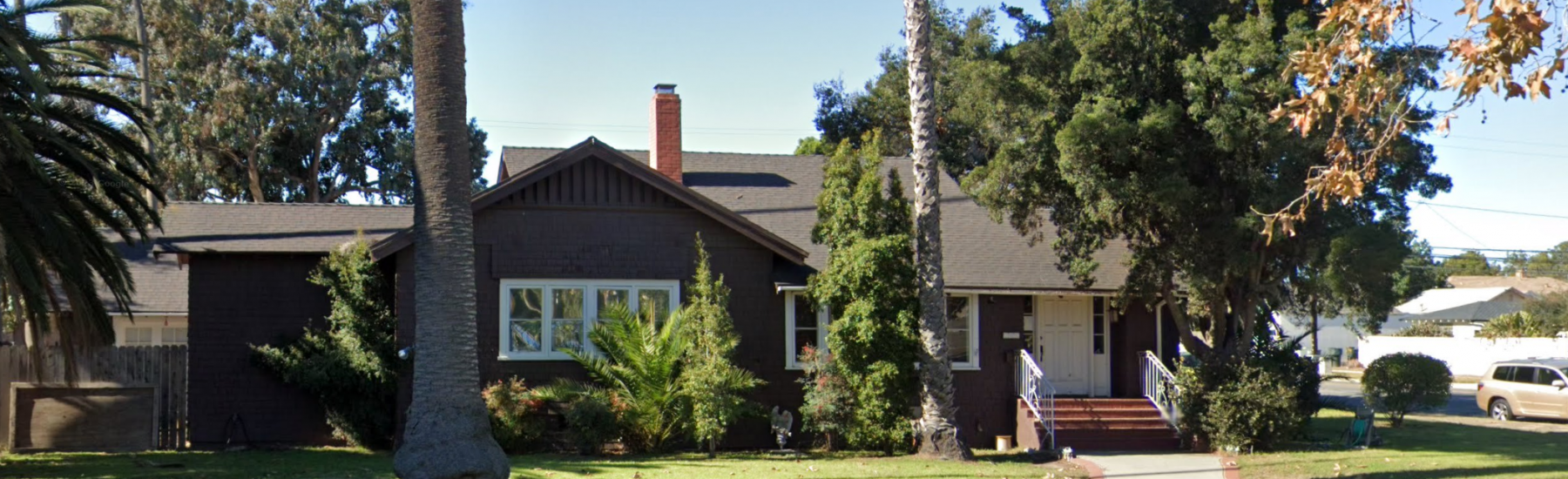 Brown single-story house with gray roof, white-framed windows, and red chimney, surrounded by trees and a well-maintained lawn. Caption: Murphy House, 2022. Credit: Google Maps.