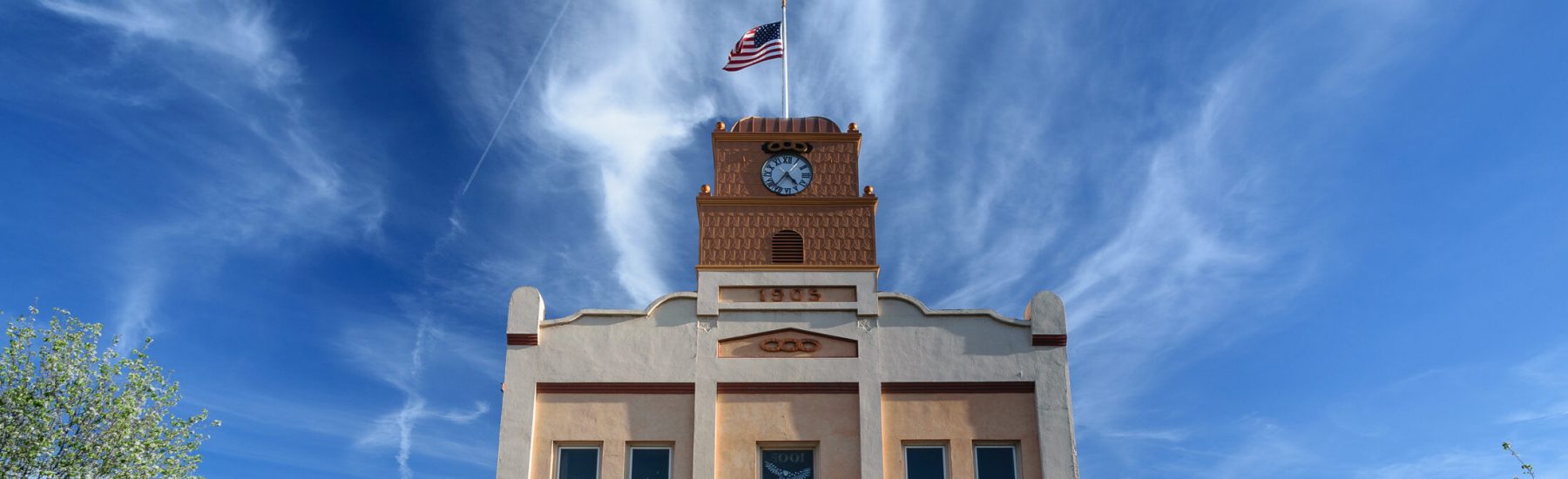 A beige building with a brown clock tower marked "1905" and "CFC", topped with an American flag. Ground floor features storefronts, benches, and flower planters under a bright blue sky.