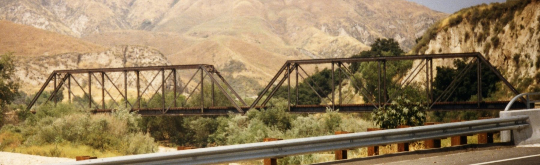 Steel truss train bridge spanning a road with guardrails, set against arid mountains and greenery, circa 1980s.