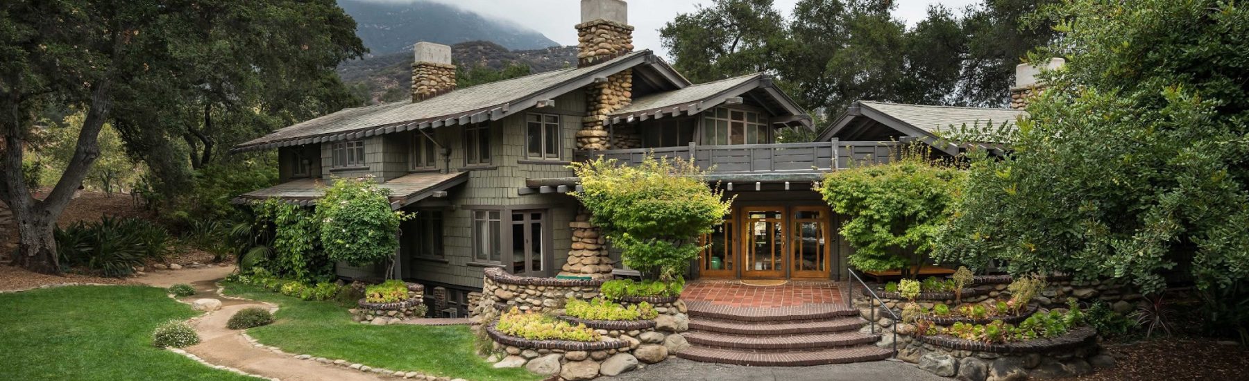 Two-story rustic house with stone chimneys, wooden doors, and a stone staircase leading to a garden, surrounded by trees and distant mountains. The Pratt House, photographed by Stephen Schafer in 2013.