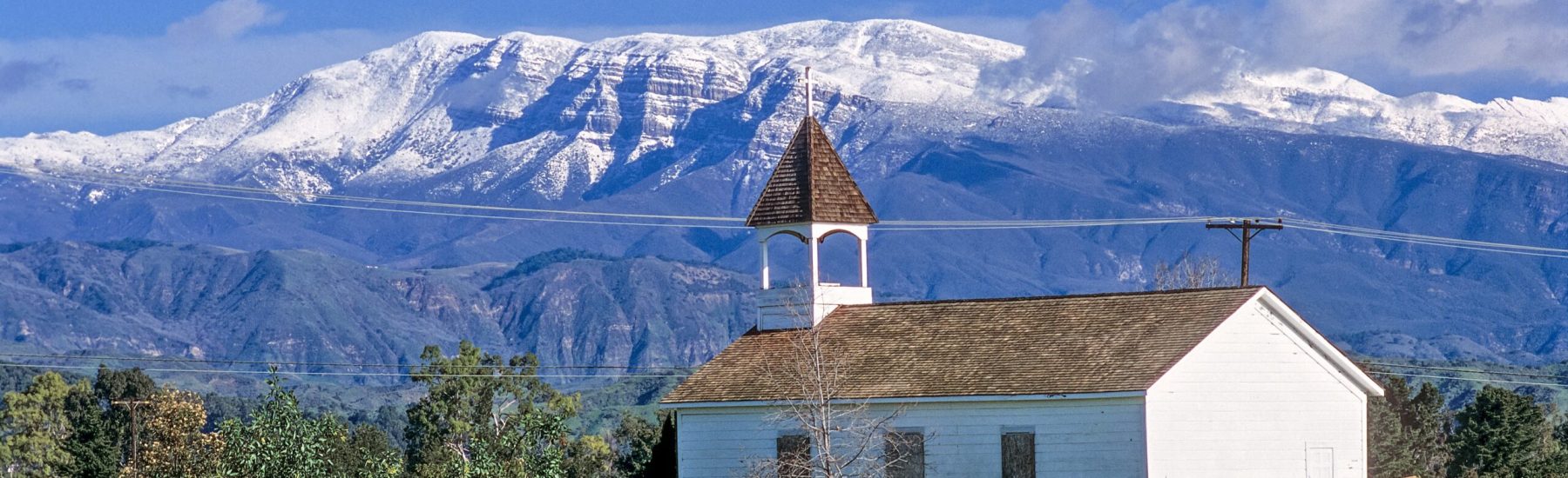 A white Sacred Heart Mission Church with a brown shingled steeple stands in a field of yellow flowers, with snow-capped mountains under a clear blue sky.