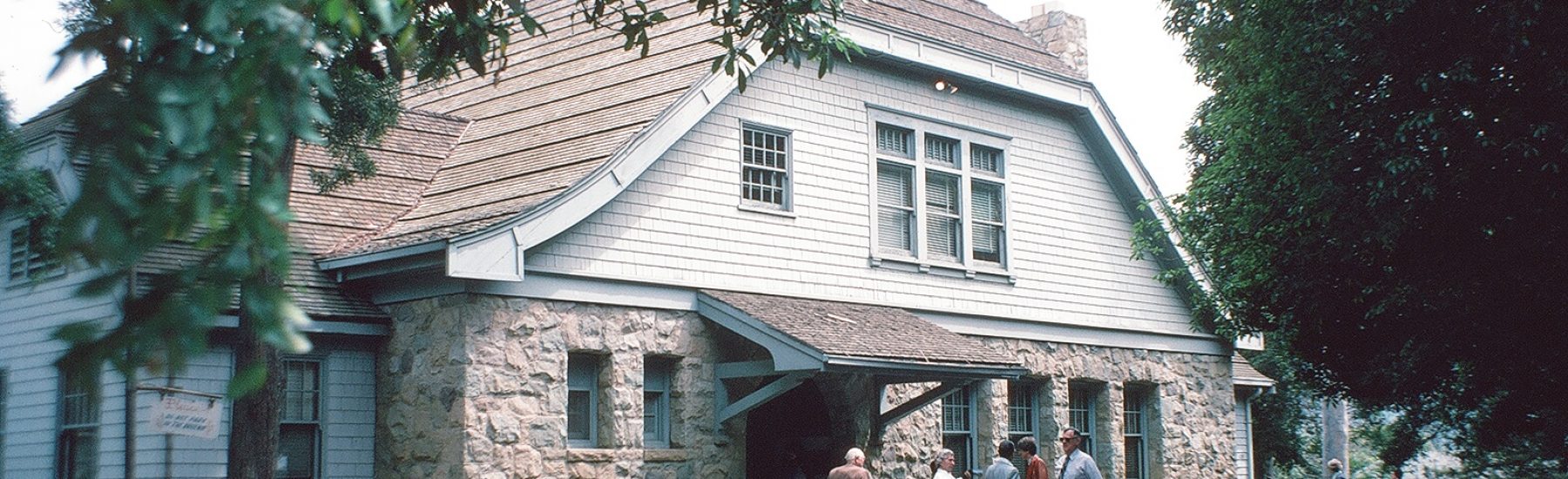 A white wooden building with a stone foundation and shingled roof, labeled 'Santa Paula Ebell Club' in 1982, features a covered porch where several people gather on a paved path.