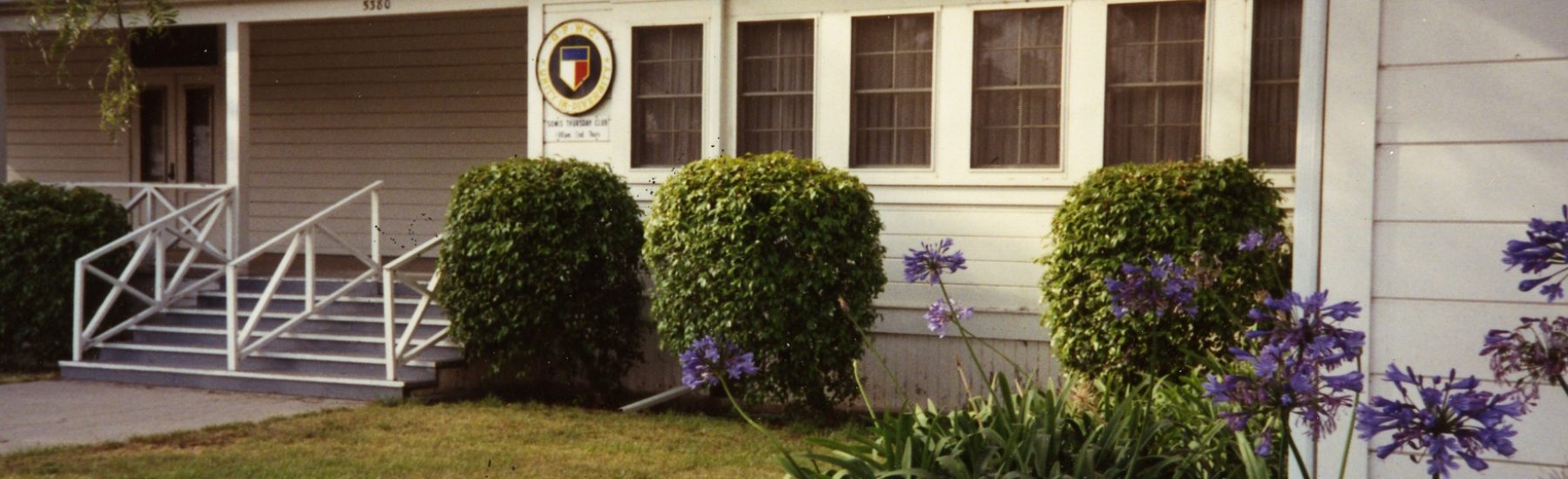 Somis Thursday Clubhouse exterior with white siding, covered porch, white railings, steps, shield emblem sign, trimmed bushes, purple agapanthus flowers, and address '5380', circa 1985.