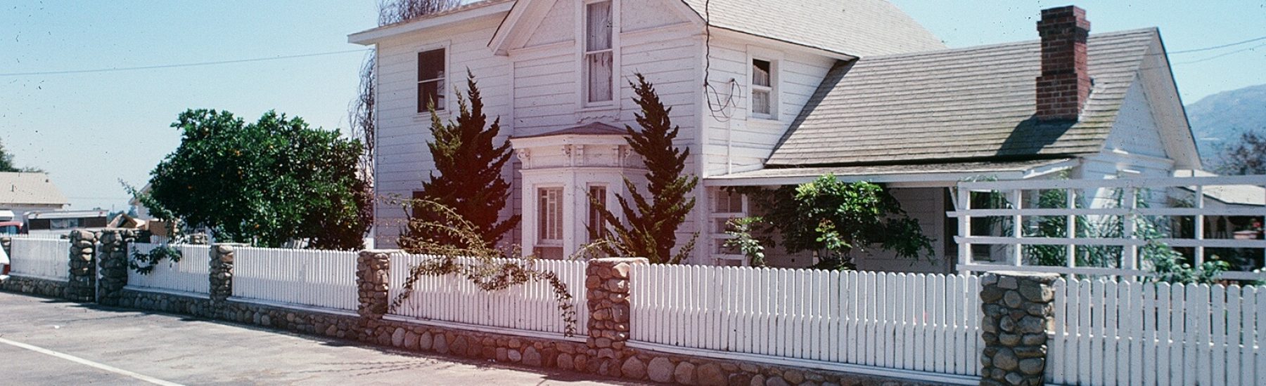 Two-story white house with gray roof and white picket fence, stone pillars, and trees on a sunny street, Tanner Homestead 1978.