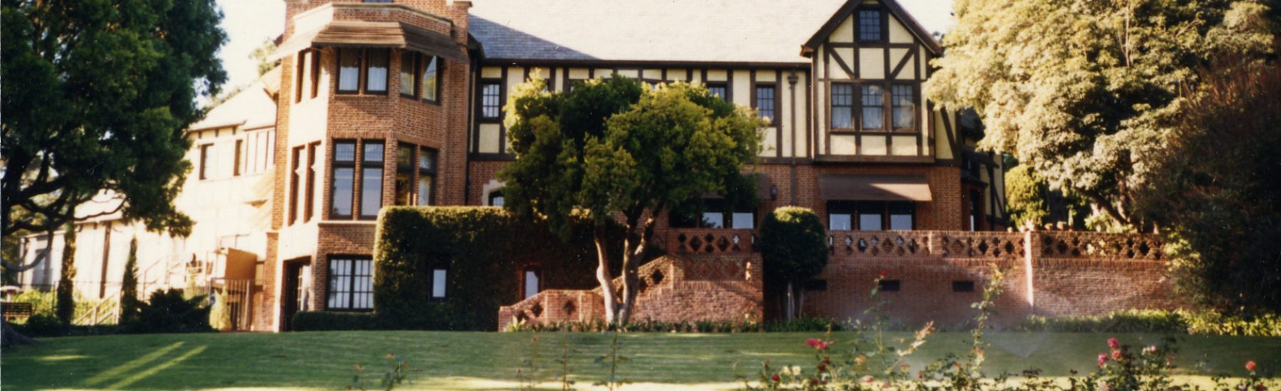 Tudor-style Teague Mansion with brick and timber framing, surrounded by trees and a green lawn with rose bushes in the foreground, photographed in 1985.