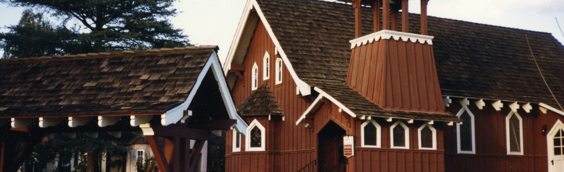 Wooden Trinity Episcopal Church with brown siding, white trim, bell tower, and arched windows, surrounded by green lawn and trees, circa 1980s.